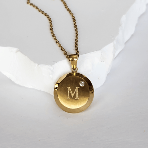 Gold necklace with a round pendant featuring the letter 'M' on a white background