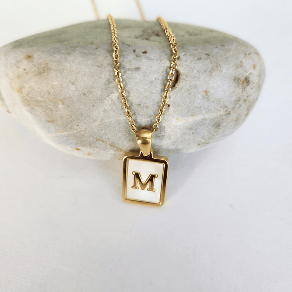 Gold necklace with a square pendant featuring the letter 'M' on a stone surface.