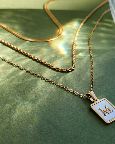 Gold necklace with a square pendant featuring the letter 'M' on a green surface.