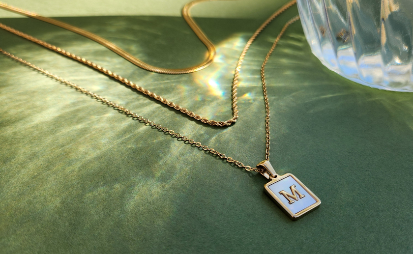 Gold necklace with a square pendant featuring the letter 'M' on a green surface.