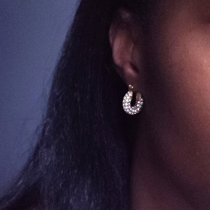A close-up of a person's ear wearing a small crescent-shaped hoop earring set with clear stones and gold-toned metal, against dark hair and a dark background.