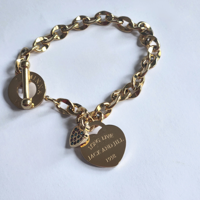 A gold-tone chain bracelet with a toggle clasp and two hanging charms: a heart-shaped charm engraved with 'LONG LIVE JACK AND JILL 1981' and a small heart-shaped charm, photographed on a plain white surface.