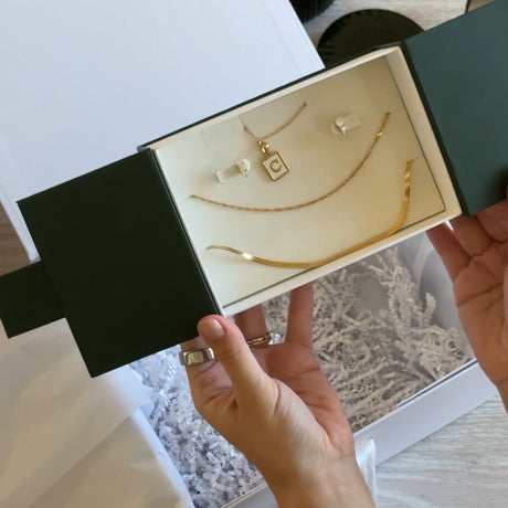 A person's hands holding open a dark green jewelry box containing gold necklaces and earrings with white pearl-like accents, set against a light wood table background.